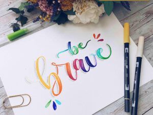 Lámina con lettering a color que dice ‘be brave’, decorada con rotuladores Tombow y flores secas en el fondo.