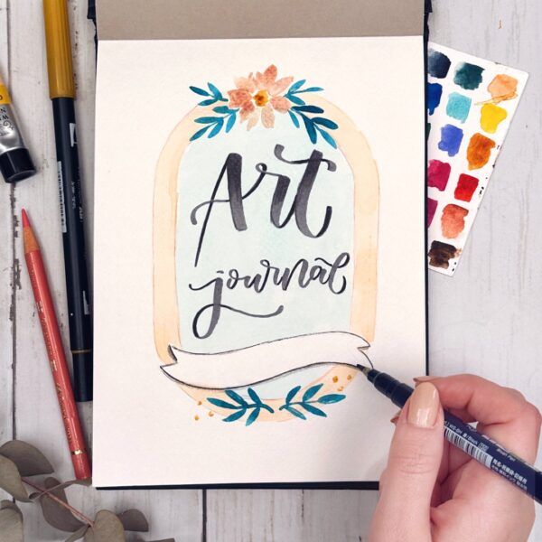 Mano dibujando en un diario de arte con la palabra 'Art journal' en caligrafía, rodeada de un diseño floral y hojas.