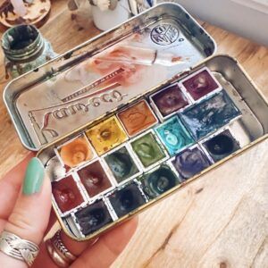 Estuche de acuarelas de viaje con colores variados para Urban Sketching, sostenido por una mano con esmalte verde.